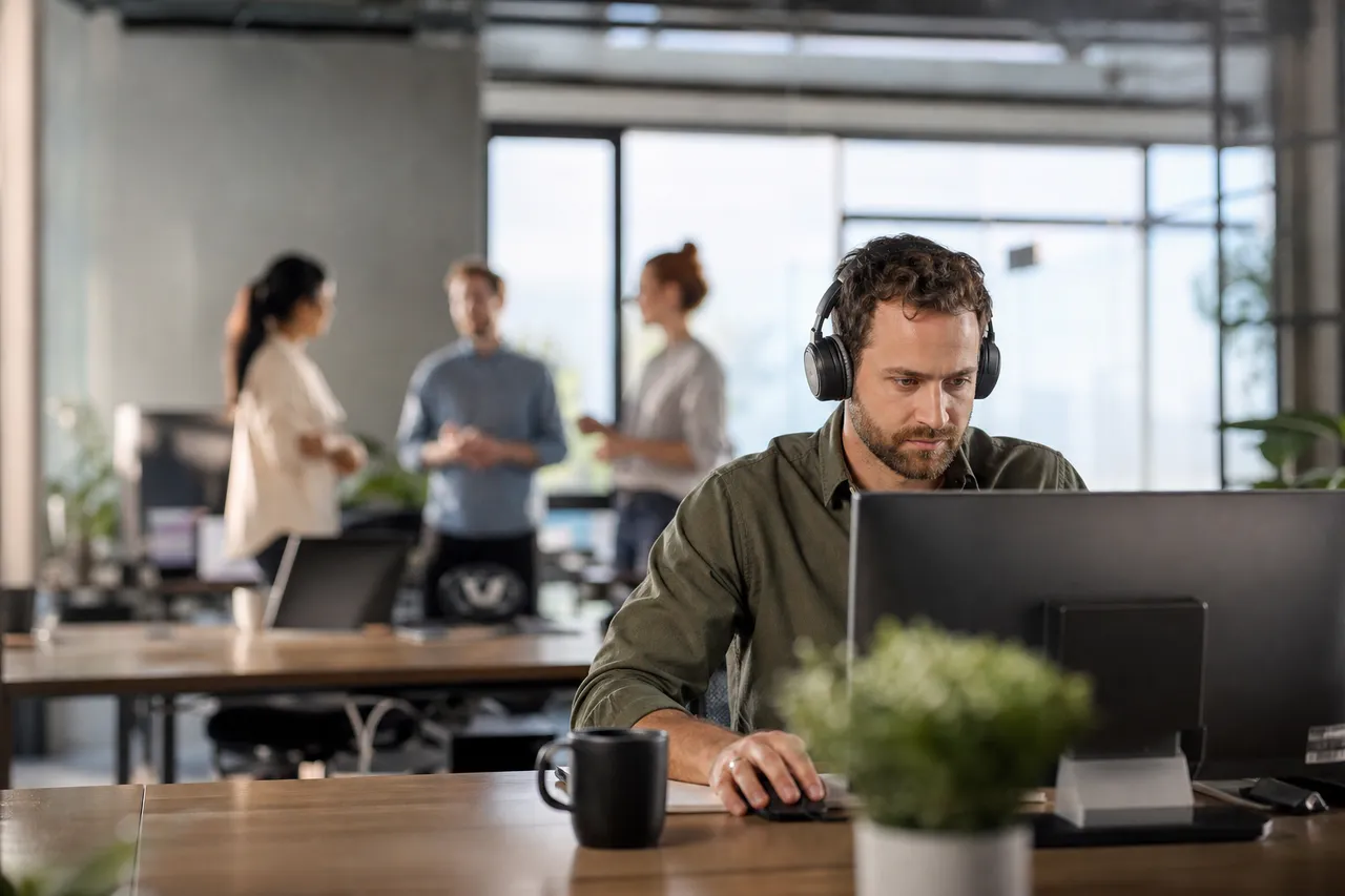Salarié avec un casque audio, concentré devant son écran dans un open space, collègues en discussion floue en arrière-plan
